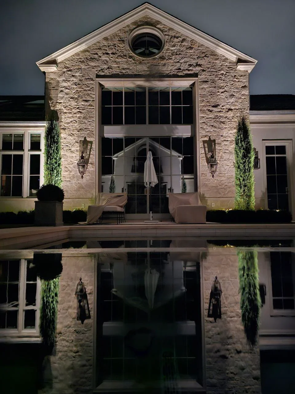 Nighttime view of illuminated stone house facade with pool reflection and landscape lighting