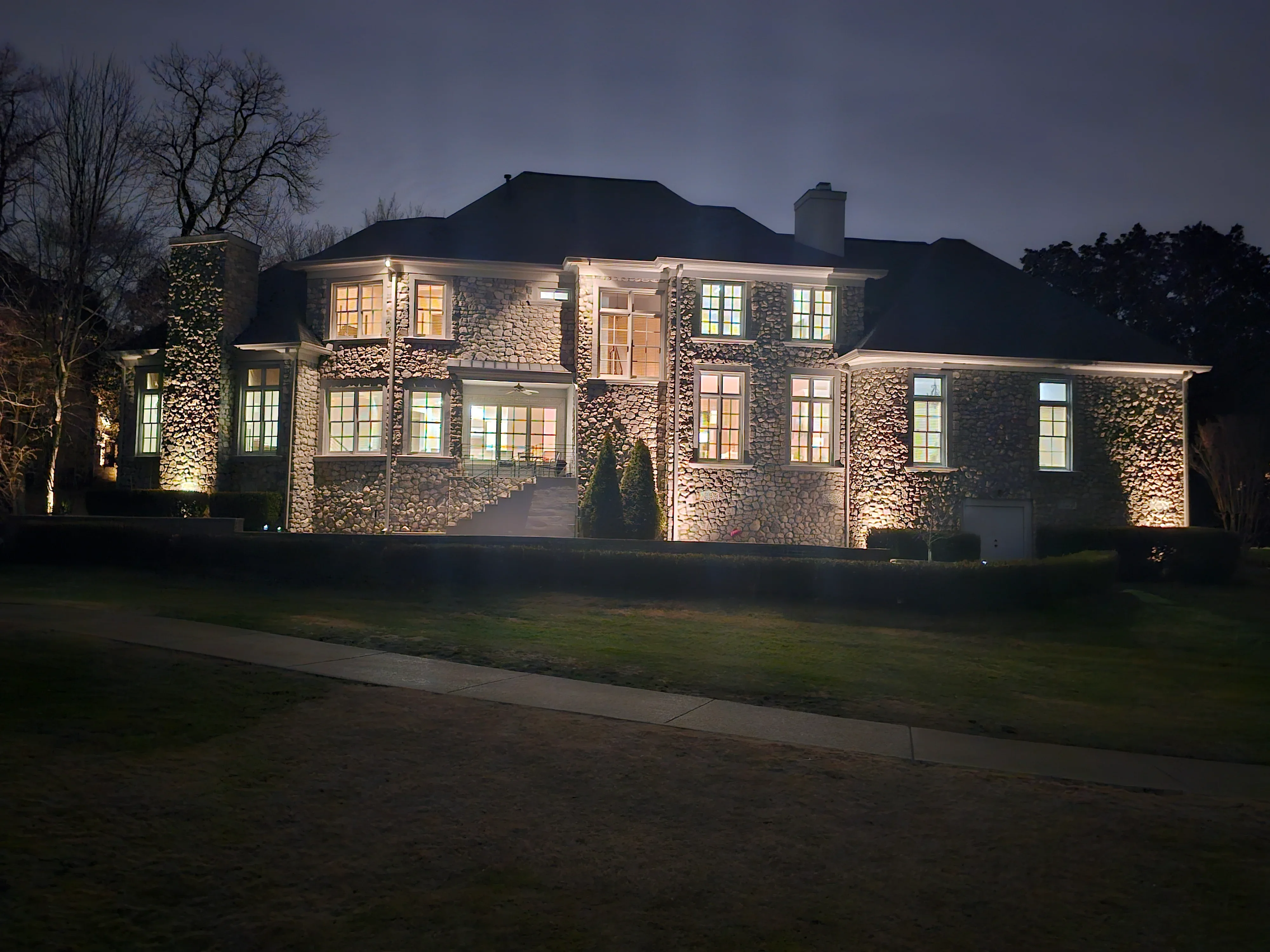 Stone estate home at dusk with warm architectural accent lighting illuminating facade and landscape features