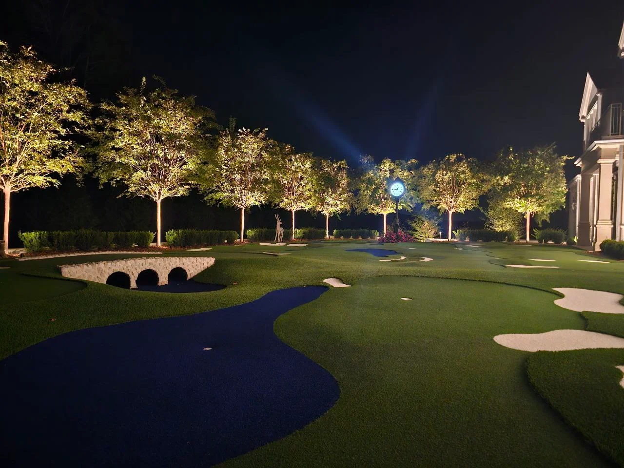 Nighttime view of illuminated residential golf course with lit trees, stone bridge, and manicured green
