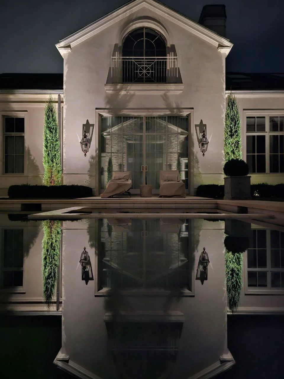 Modern farmhouse exterior at night with uplighting on white facade and entry sconces