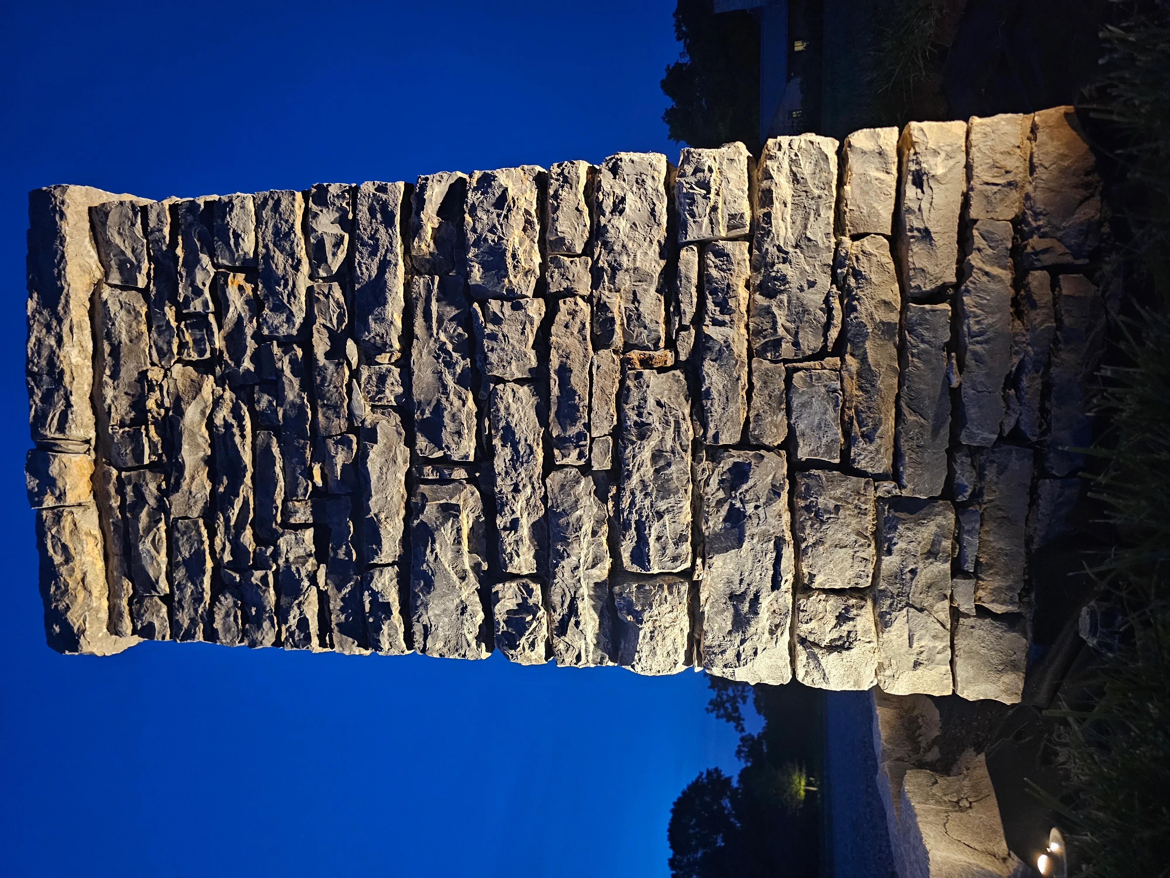 Warm uplighting on textured stone retaining wall at dusk with clear blue sky